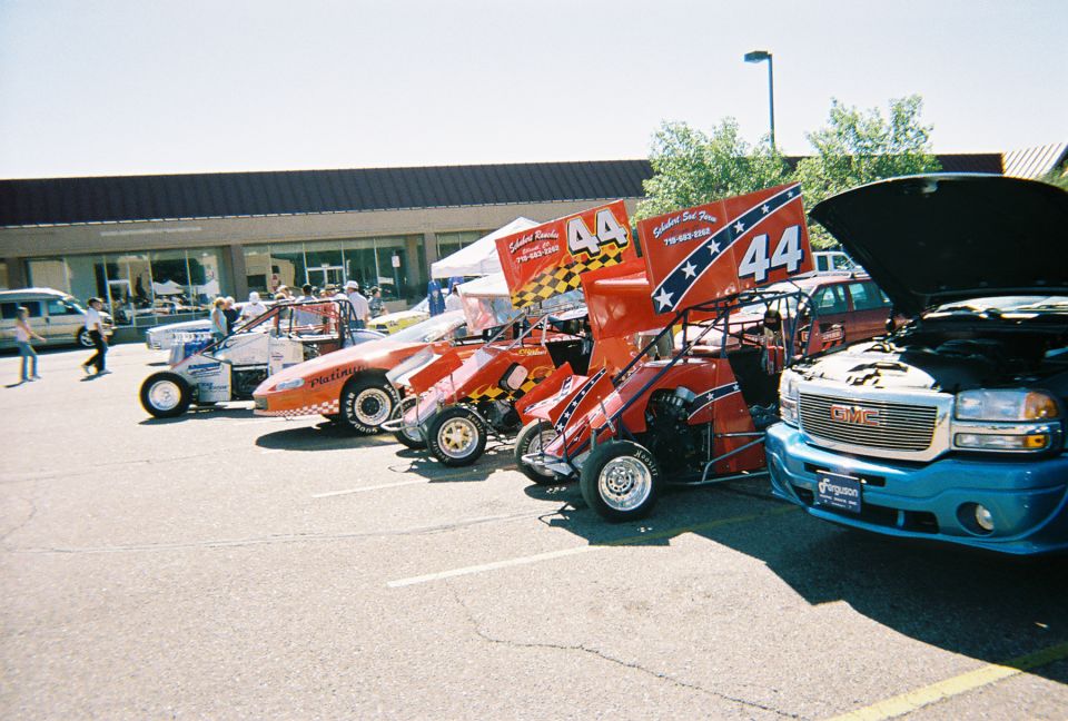Row of red number 44 sprint cars parked beside a blue GMC pickup at an outdoor show