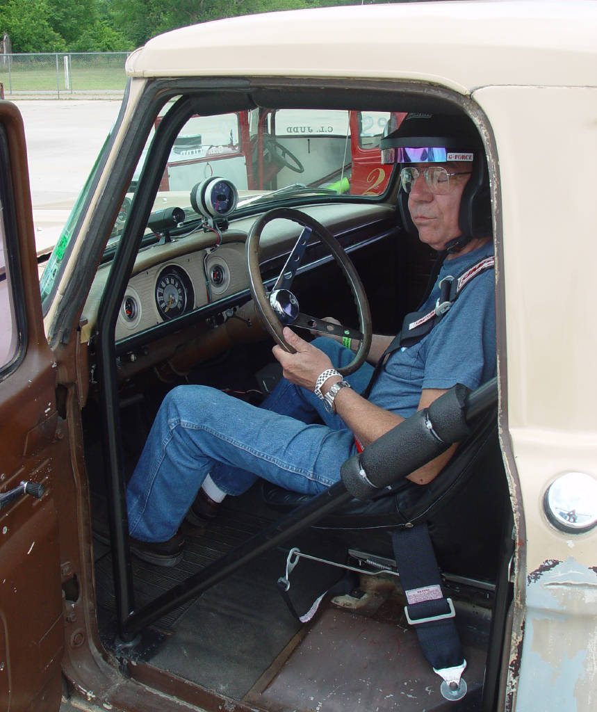 Man wearing a racing helmet and goggles sits behind the steering wheel of a vintage pickup truck. The interior shows a roll cage and racing harness