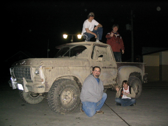 Muddy older Ford pickup at night with four people posed on and around it, 'HELP' written in mud