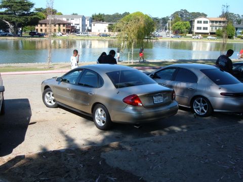 1995 Ford Taurus - Two sedans are parked near a lake with people in the background.