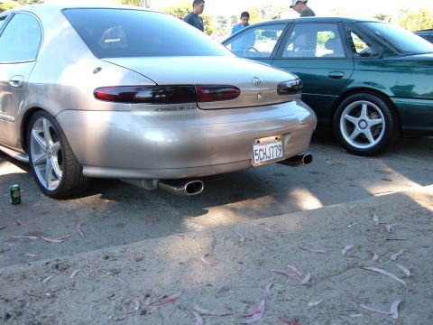1995 Ford Taurus - Rear view of a gold sedan with dual exhaust pipes and aftermarket wheels, parked next to a green sedan.