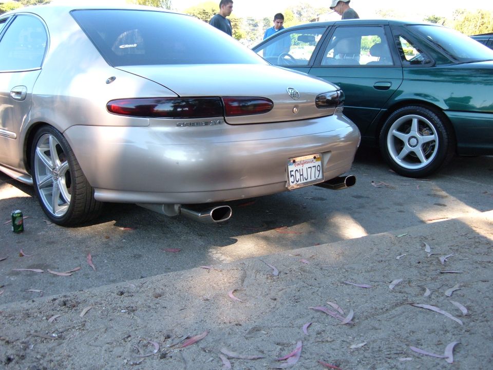Gold sedan's rear end, dual exhaust, and aftermarket wheels. A green sedan is parked to the right