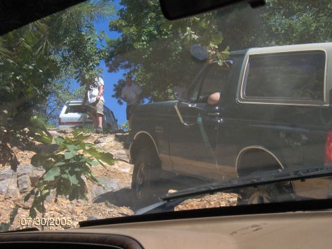 1978 Ford Bronco - Two SUVs navigate a rocky trail on a sunny day, with one vehicle partially obscured by foliage.