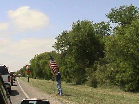 1987 Ford Bronco - People wave American flags from the roadside as traffic passes by on a sunny day.