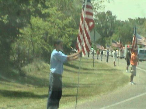1987 Ford Bronco - A group of people are holding American flags in a line along a grassy area next to a road.