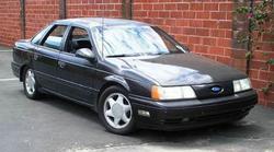 1991 Ford Taurus - This dark sedan is parked outside a brick building with some greenery on the right.