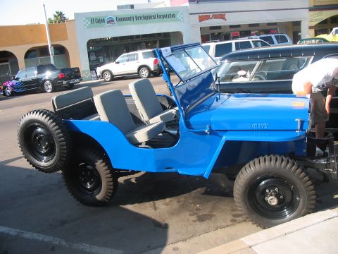 1986 Ford Bronco - A blue Willys Jeep is parked on a street, with other vehicles and buildings in the background.