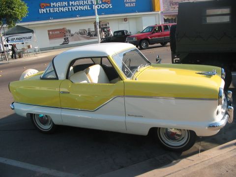1986 Ford Bronco - A two-tone yellow and white Nash Metropolitan is parked on a street, with other vehicles and buildings in the background.