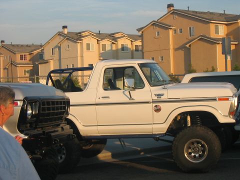 1986 Ford Bronco - A lifted white Ford Bronco is parked next to another vehicle, with apartment buildings in the background.