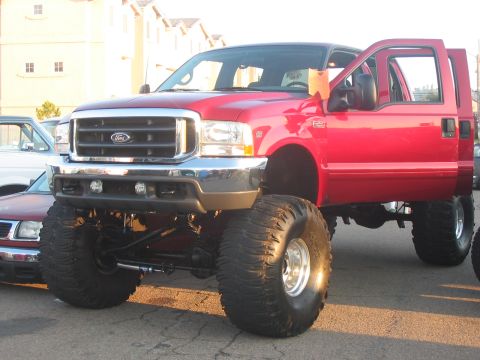 1986 Ford Bronco - Lifted red Ford F-250 Super Duty truck with large off-road tires and front doors open.