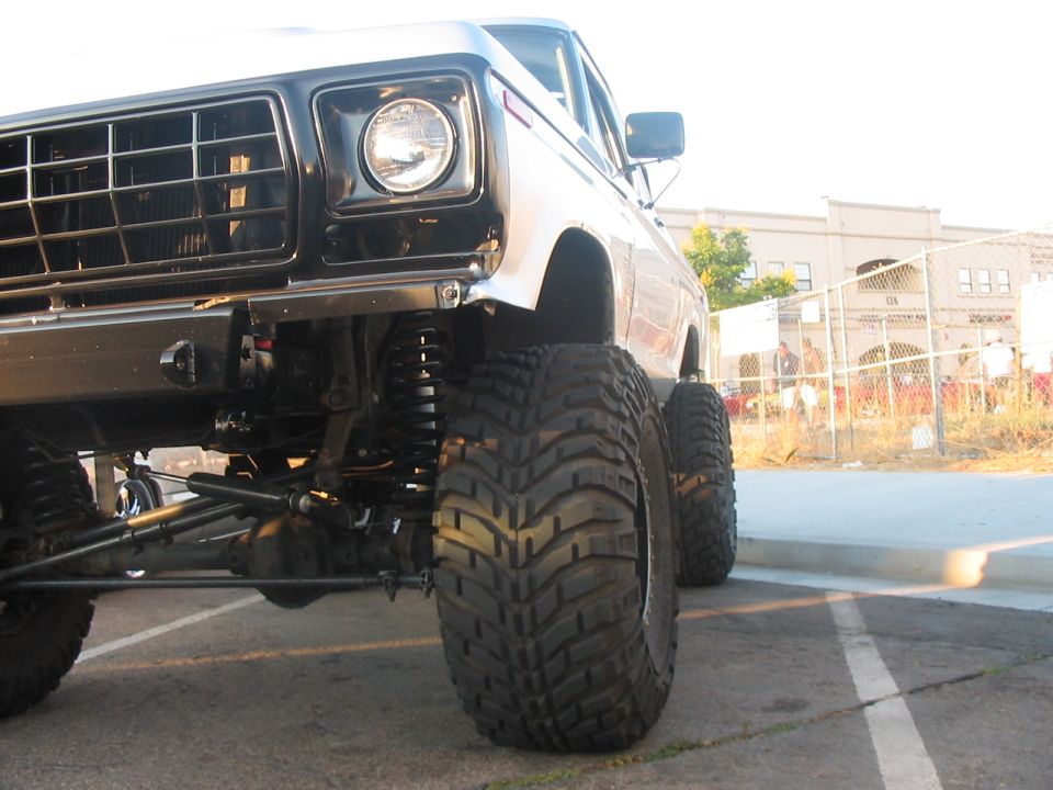 Front corner of a lifted pickup truck with large off-road tires. Grille and headlight visible