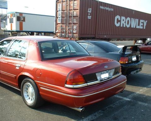 1999 Ford Crown Victoria - Rear view of a red 1999 Ford Crown Victoria, parked in a lot with other vehicles and a shipping container.