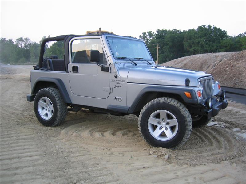 Silver Jeep Wrangler Sport with top removed parked on sand at gravel pit