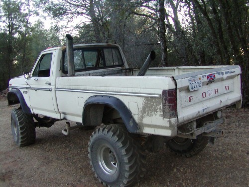 Lifted white Ford pickup truck, rear view, with large exhaust pipes and custom wheels. License plate is visible