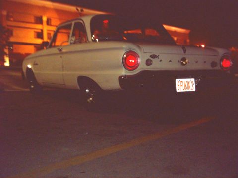 1962 Ford Falcon - A white 1962 Ford Falcon sedan is seen from the rear at night, illuminated by building lights.