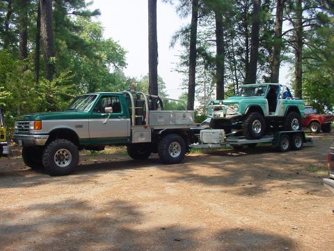 1969 Ford Bronco - A lifted Ford Bronco is transported on a trailer by a large Ford F-Series truck, with a red vehicle visible in the background.