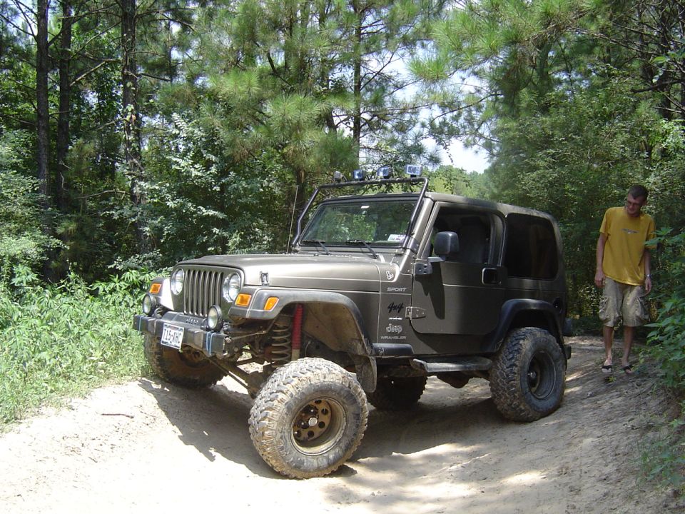 Olive Jeep Wrangler Sport, lifted on mud tires with light bar, on a dirt trail; man standing at right