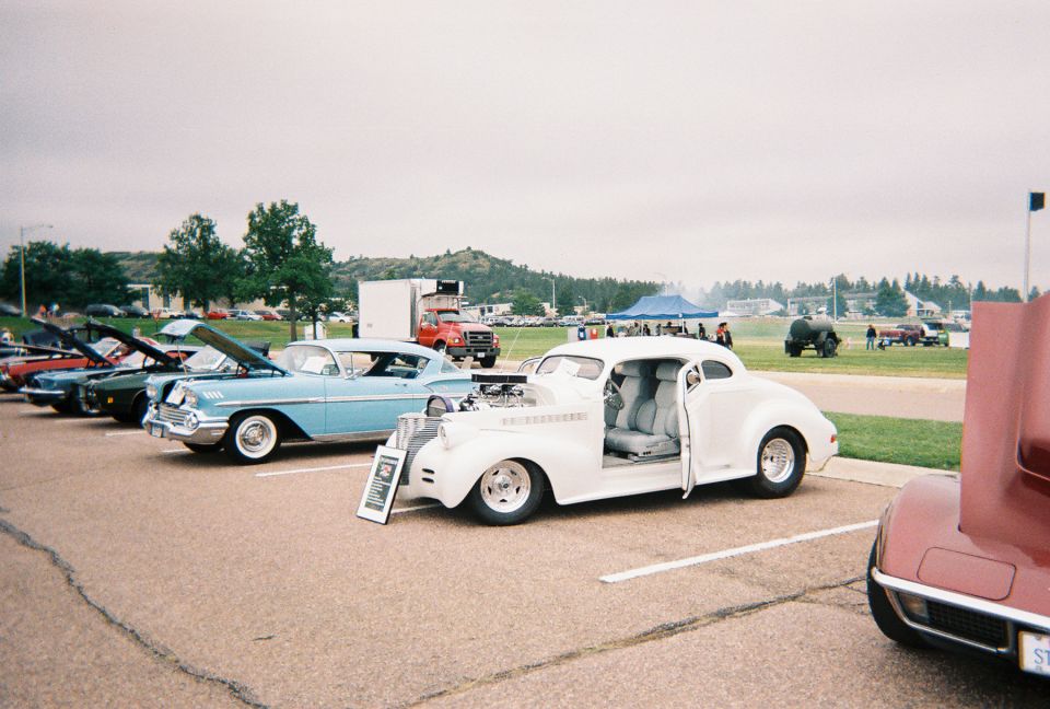 White chopped 1930s hot rod coupe with exposed engine and open door at an outdoor car show