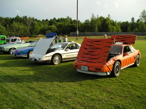 1984 Pontiac Fiero - Several cars are parked on a grassy field, including a white Pontiac Fiero and an orange one with tiger stripes.