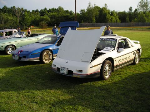 1984 Pontiac Fiero - A white 1984 Pontiac Fiero, labeled as an official race car, is displayed with its hood open next to a blue Fiero.