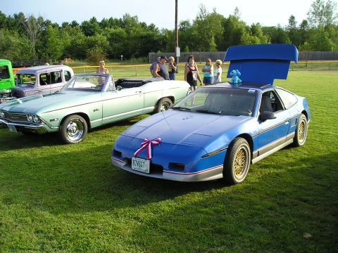 1984 Pontiac Fiero - Two cars are parked on a grassy field, with trees in the background and people standing around.