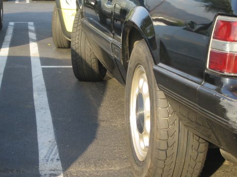 1985 Ford Mustang - A black car's rear tire and fender are visible, parked next to a yellow vehicle.