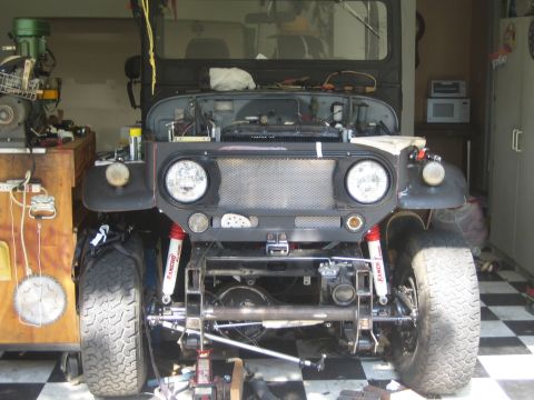 1990 Ford Bronco - stripped black off-road vehicle sits mid-build in a checkered-floor garage, exposing its grille, lights, and front axle.