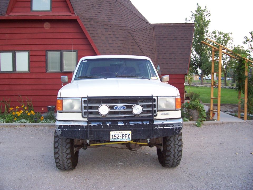 Front view of a white Ford Bronco with a bull bar and off-road tires parked in front of a red house