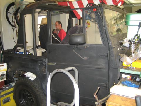 1990 Ford Bronco - man sits at the wheel of a stripped black open-top 4x4 under construction in a cluttered garage with a flag overhead.