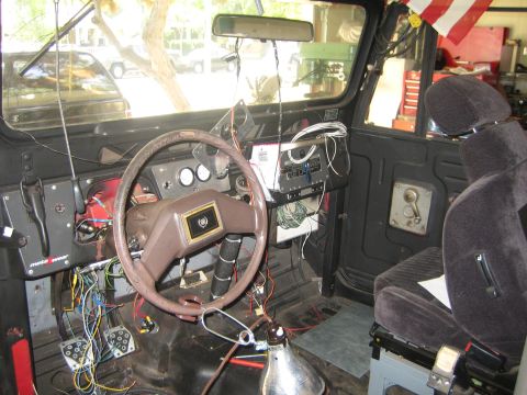 1990 Ford Bronco - Interior of a vehicle under repair, with a worn wrapped steering wheel, dangling wires under the dash and a gray cloth bucket seat.