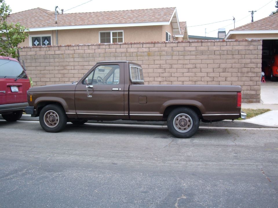 Side view of a brown 1985 Ford Ranger pickup truck parked next to a red SUV