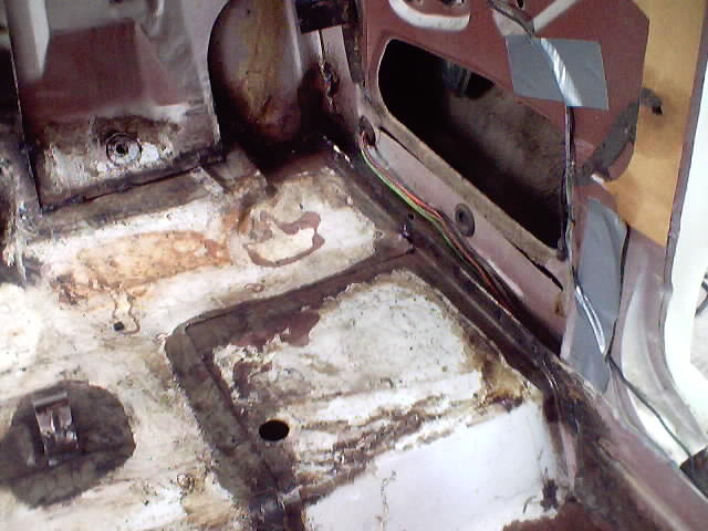 Interior floor pan of a car showing rust and dirt, with wiring and a section of a maroon panel. Supermotors.net logo visible