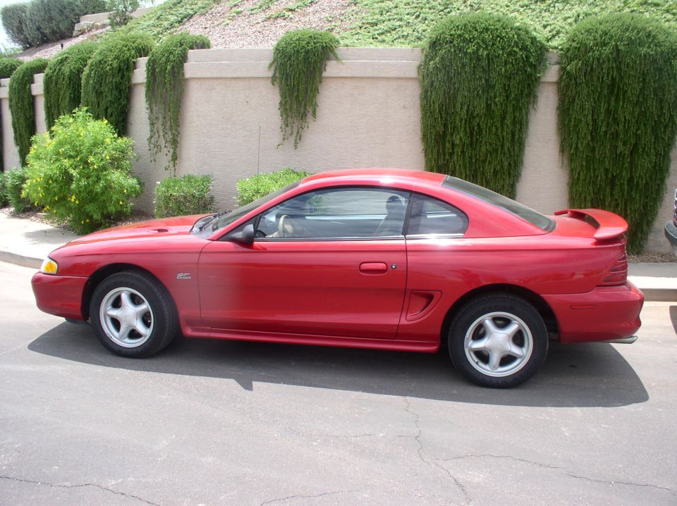 Red coupe parked on asphalt, side view. Features a spoiler and five-spoke wheels