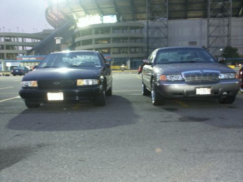 2001 Mercury Grand Marquis - Two sedans, one a gray 2001 Mercury Grand Marquis, are parked in a lot with a stadium in the background.