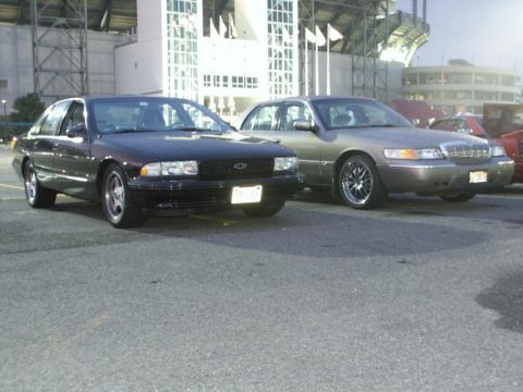 2001 Mercury Grand Marquis - A tan 2001 Mercury Grand Marquis is parked next to a black Chevrolet sedan in front of a stadium.