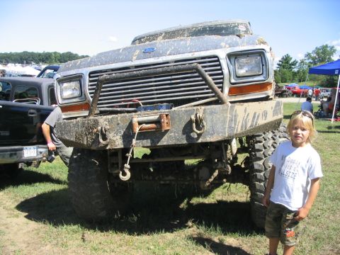 1986 Ford Bronco - A child poses next to a heavily modified, mud-splattered Ford truck from the 1980s at an outdoor event.