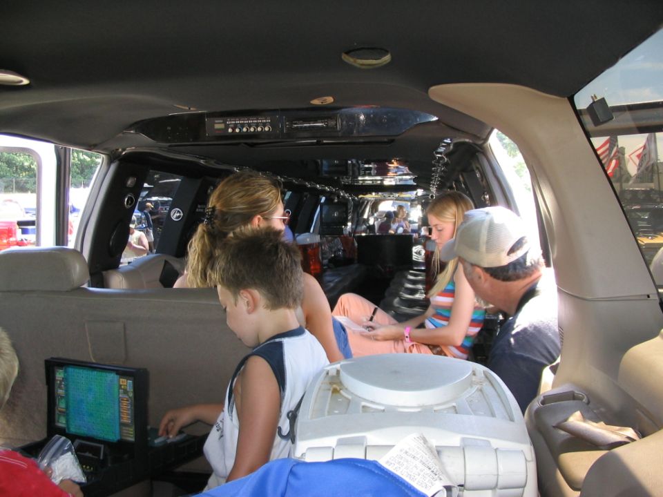 Family inside a modified SUV or van, looking at a screen and sitting on seats. A cooler is in the foreground