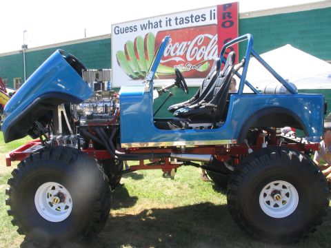 1986 Ford Bronco - This modified off-road vehicle has a prominent V8 engine and large tires, with a Coca-Cola billboard in the background.