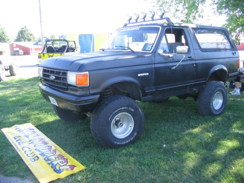 1986 Ford Bronco - A lifted black 1986 Ford Bronco is parked on grass, with a yellow "4x4 Club" banner in front and another vehicle in the background.