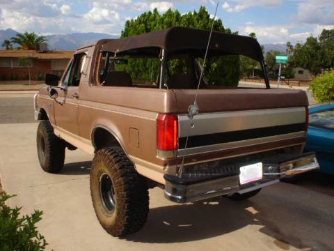 1989 Ford Bronco - This brown soft-top SUV features a lifted suspension and large off-road tires, seen from the rear.