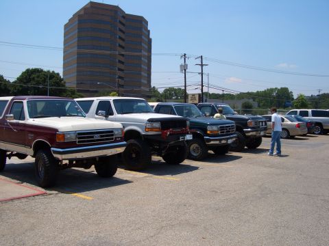 1990 Ford Bronco - A lineup of lifted Ford trucks and Broncos are parked in a lot on a sunny day, with a tall office building in the background.