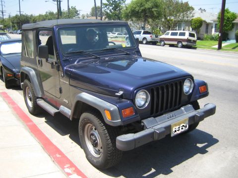 1990 Ford Bronco - dark blue Jeep Wrangler with a black soft top sits parked at a red-painted curb along a sunny suburban street.