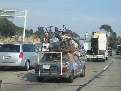 1990 Ford Bronco - battered blue hatchback piled high with furniture and bicycles on a tall roof rack crawls through freeway traffic beside a box trailer.