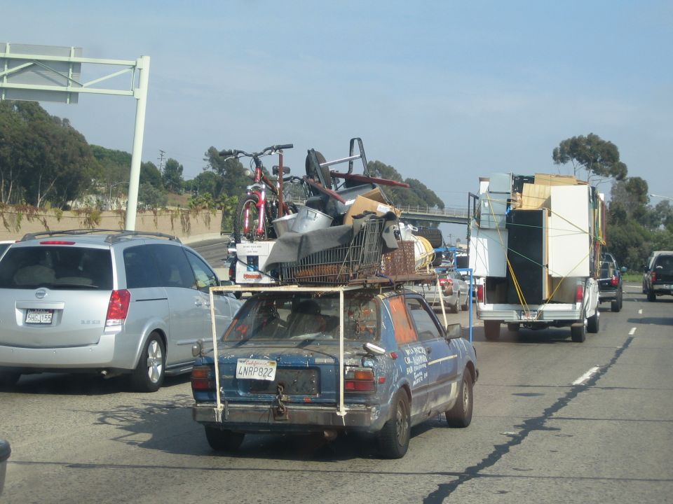 Blue hatchback overloaded with furniture and bikes on a roof rack in freeway traffic