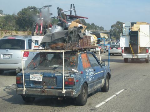 1990 Ford Bronco - Rear view of an overloaded blue hatchback hauling furniture, a shopping cart and bikes on its roof rack through dense freeway traffic.