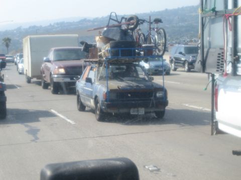 1990 Ford Bronco - small blue car heaped with chairs, bikes and household goods on a roof rack sits in slow traffic among other vehicles.
