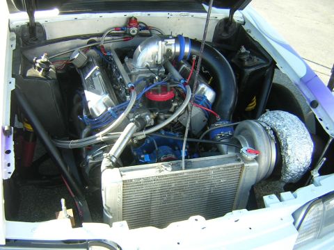 1998 Ford Crown Victoria - Engine bay of a white car fitted with a twin-turbo V8, polished intake, braided blue lines and a front-mounted aluminum cooler.
