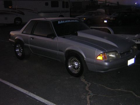 1998 Ford Crown Victoria - silver Fox-body Ford Mustang LX coupe with chrome drag-style wheels parked at dusk near RVs and other vehicles.