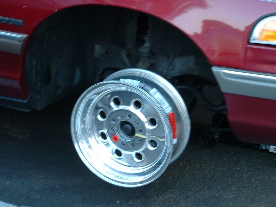 Polished aluminum wheel leaning against the raised front of a dark red sedan on a jack.