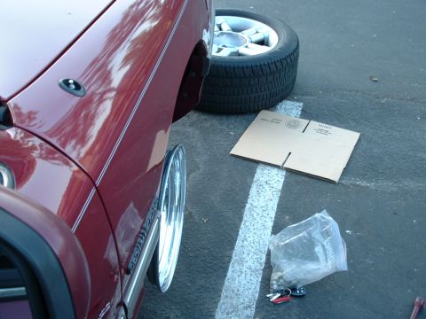 1992 Ford Crown Victoria - Wheel swap in progress in a parking lot: a dark red sedan sits with its front wheel removed and the bare hub exposed, the stock lace-style…